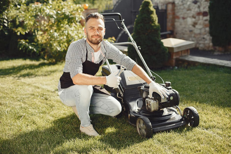 A man with a tidy beard and short brown hair, dressed in a checkered shirt, black apron, and light blue jeans, is kneeling on a well-maintained grassy lawn while holding a lawnmower with both hands. T